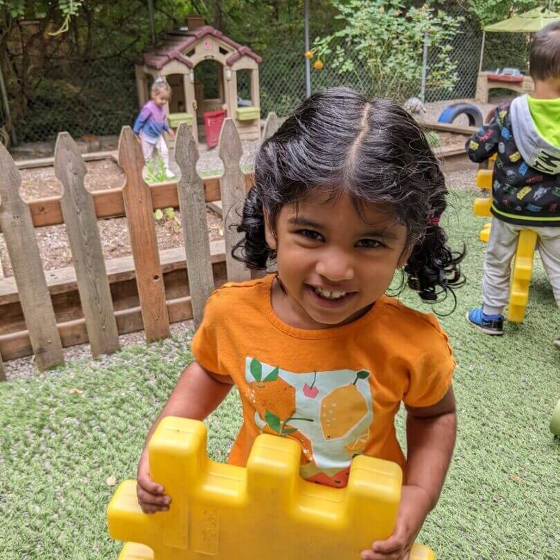 Smiling child holding large yellow block outside.